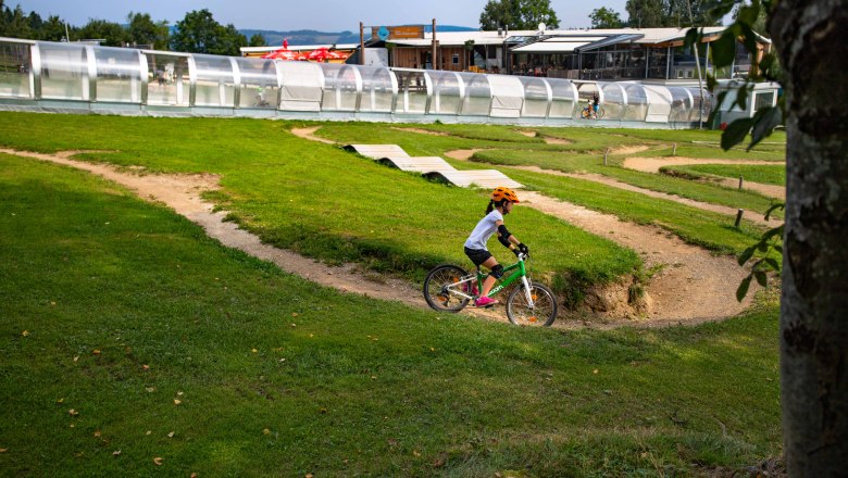 Ein Kind mit orangem Helm fährt auf einem grünen Fahrrad durch einen Mini-Bikepark mit Hügeln und Kurven.