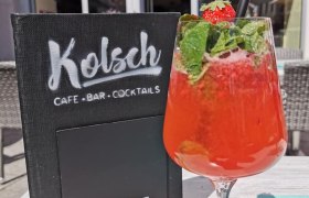 Ein roter Cocktail mit Minze und Erdbeere vor einem Caf&eacute; mit Schild 'K&ouml;lsch Caf&eacute; Bar Cocktails'.