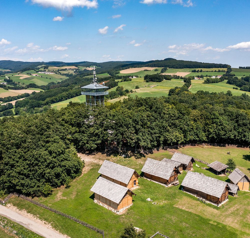 Luftansicht des Keltendorf Schwarzenbach mit den Häusern auf der Wiese und dem Aussichtsturm umgeben von Wald, Feldern und den Hügeln der Buckligen Welt.