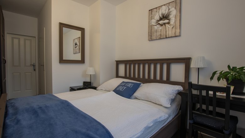 Cozy double room with wooden bed, blue ceiling and decorative cushions. A mirror and a flower picture on the wall, next to it a desk with a plant.