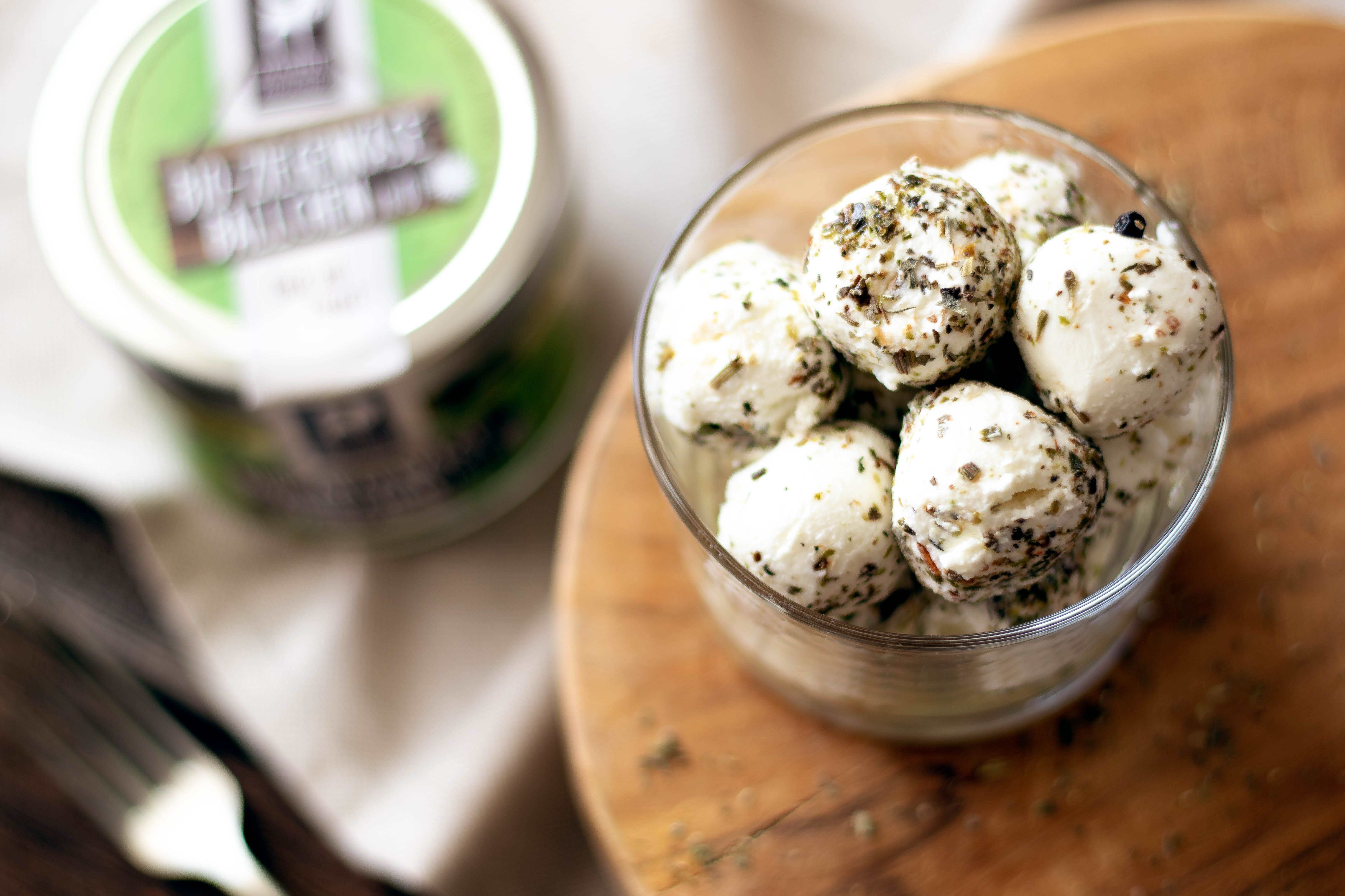 Ein Glas gefüllt mit Ziegenkäsebällchen, bestreut mit Kräutern, auf einem runden Holzbrett. Im Hintergrund ein weiteres Glas Ziegenkäsebällchen mit grünem Etikett.