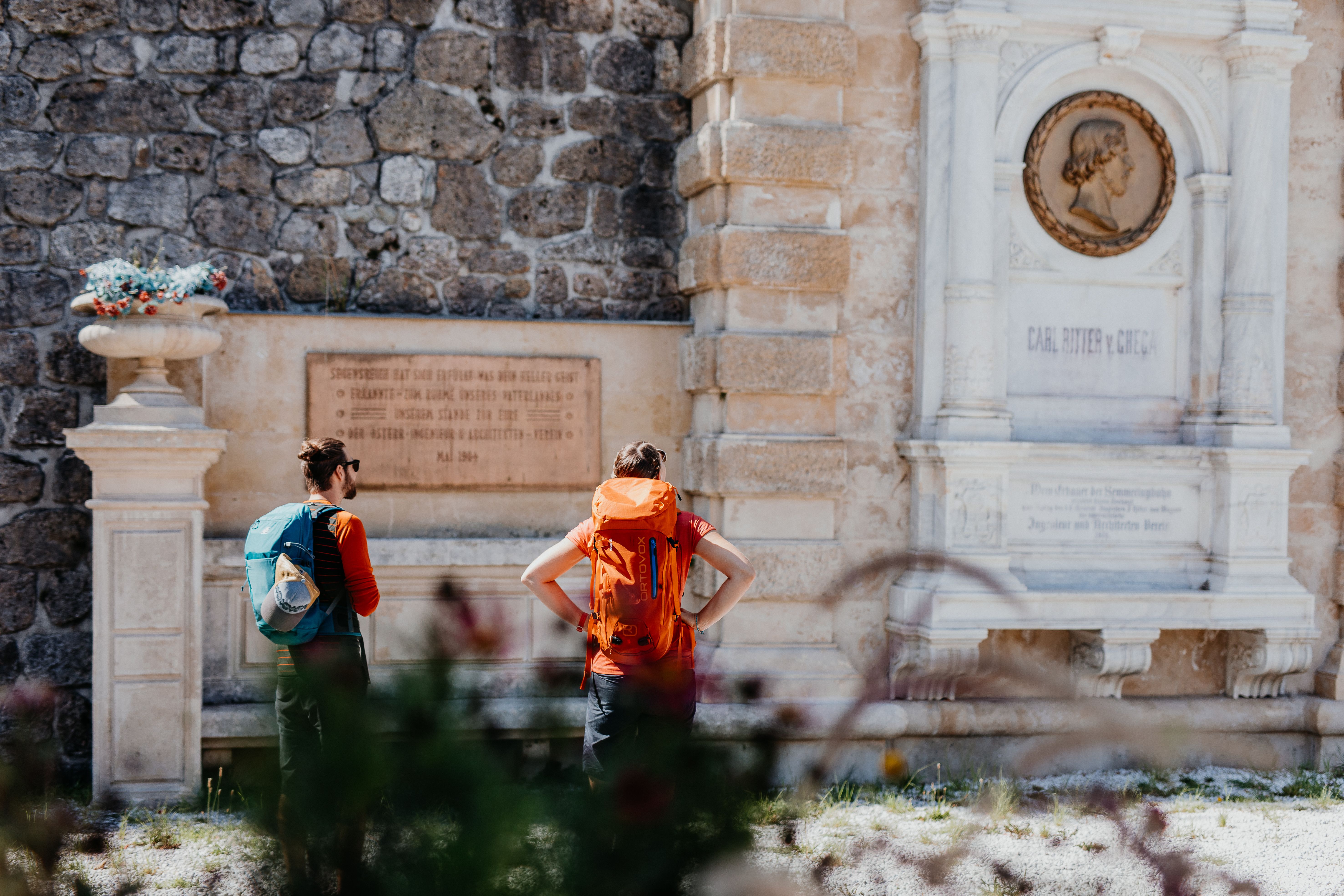 Zwei Wanderer stehen fasziniert vor einer historischen Gedenkstätte, umgeben von der majestätischen Kulisse der Wiener Alpen. Die warme Sonne strahlt auf die alten Steine, während die frische Bergluft die Sinne belebt. Hier, wo Geschichte und Natur aufeinandertreffen, wird jeder Schritt zu einem unvergesslichen Erlebnis.