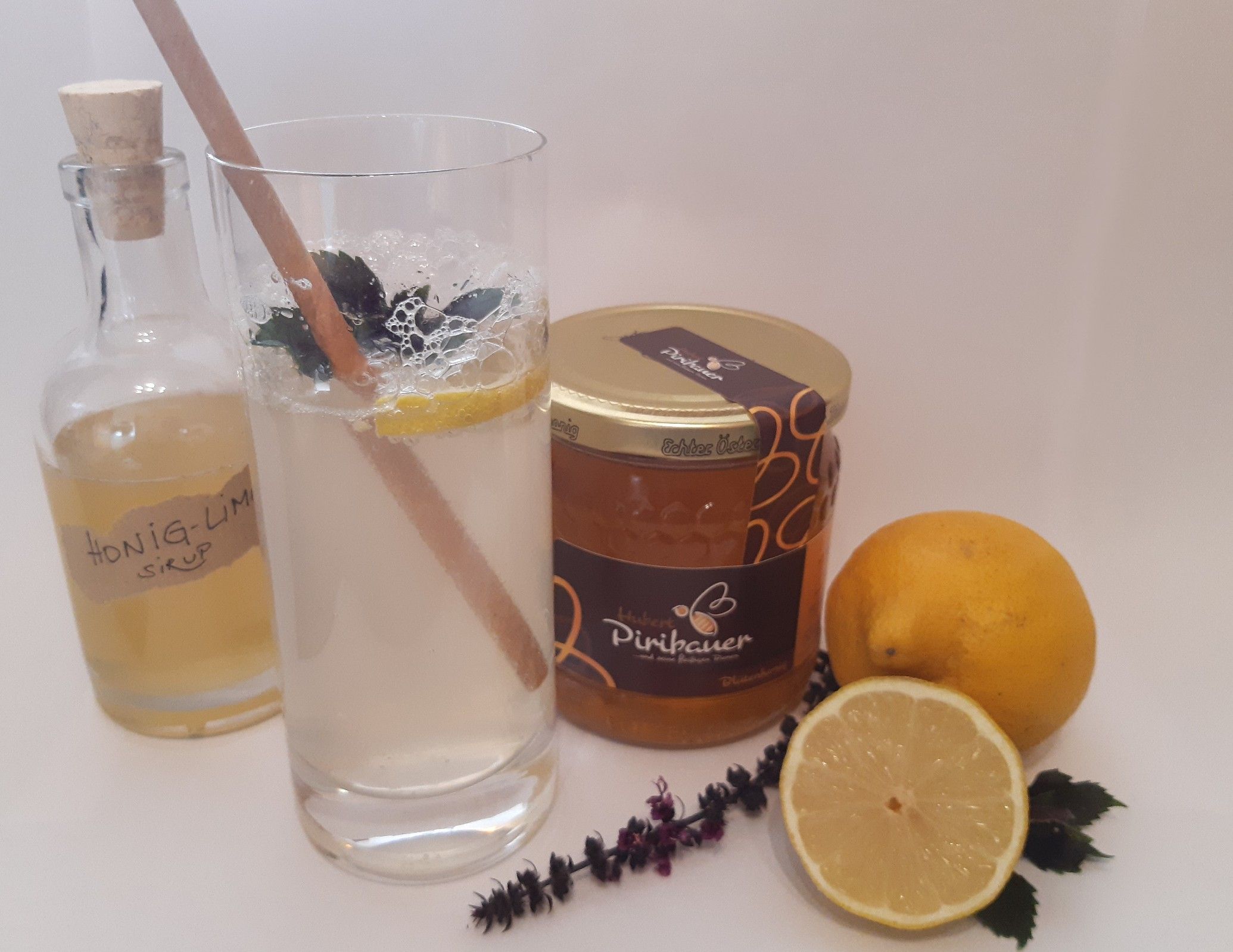 Ein Glas Honiglimonade mit Minze und Zitrone, daneben Honigglas, Zitronen, Kräuter und Sirupflasche.