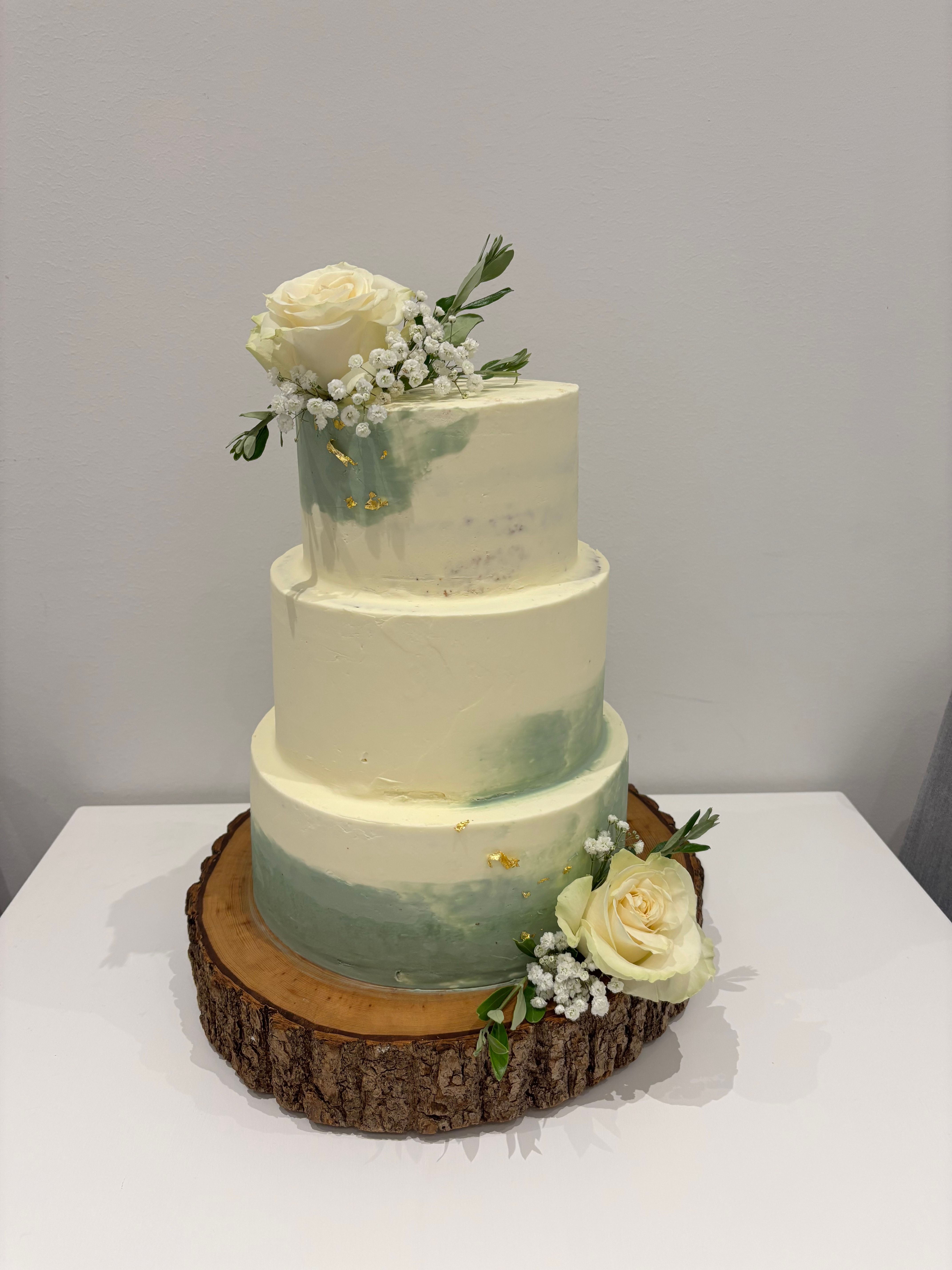 Zweistöckige Torte mit cremefarbenem und grünem Farbverlauf, dekoriert mit weißen Rosen, Schleierkraut und goldenen Akzenten auf einer Holzscheibe.