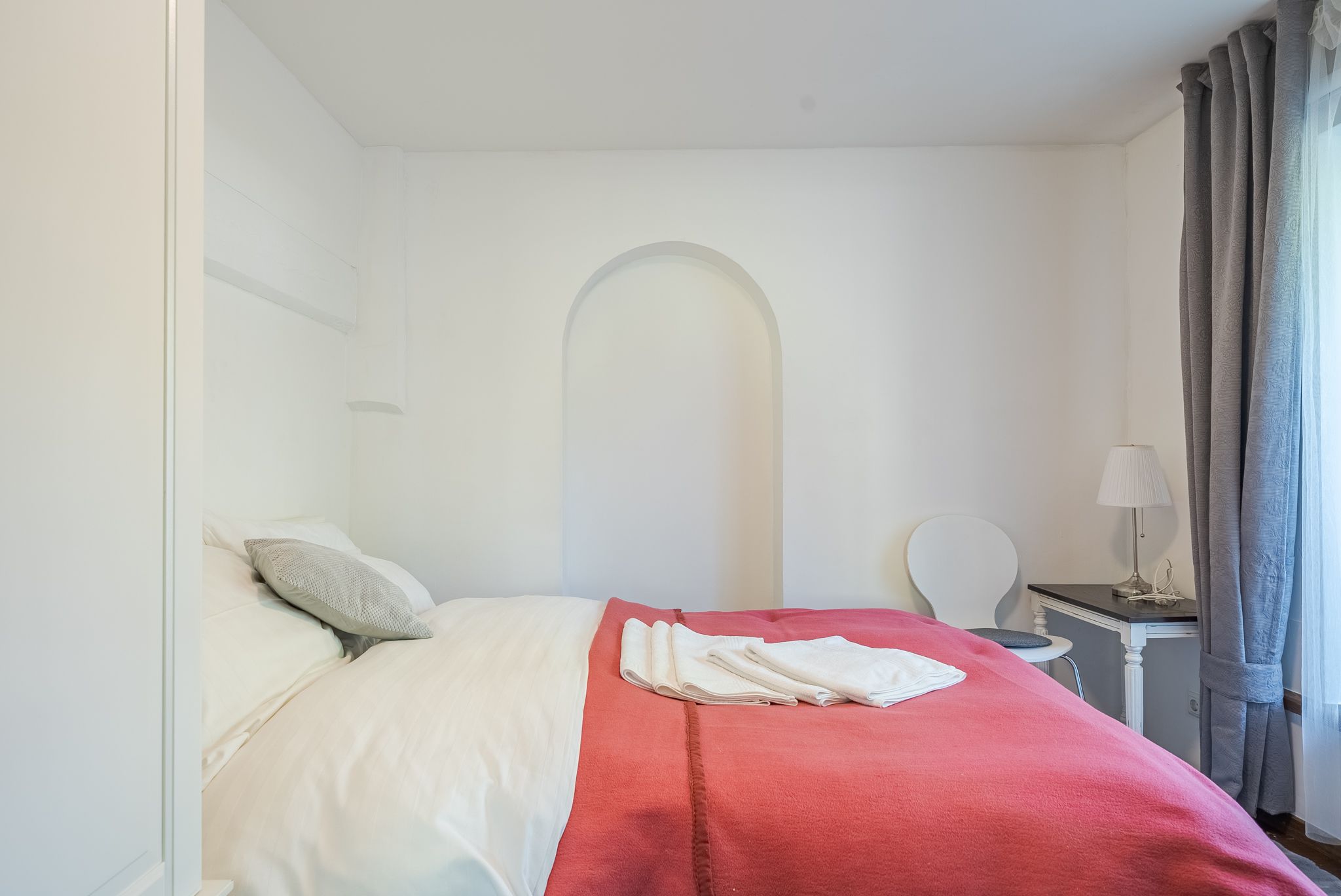Ein gemütliches Schlafzimmer mit einem Doppelbett, weißen Wänden und einem roten Überwurf. Ein kleiner Tisch mit Lampe steht neben dem Bett.
