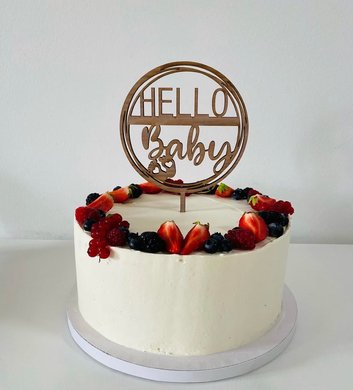 Runde, weiß eingestrichene Torte mit Beerenkranz und Holzstecker „HELLO Baby“ auf weißem Hintergrund.