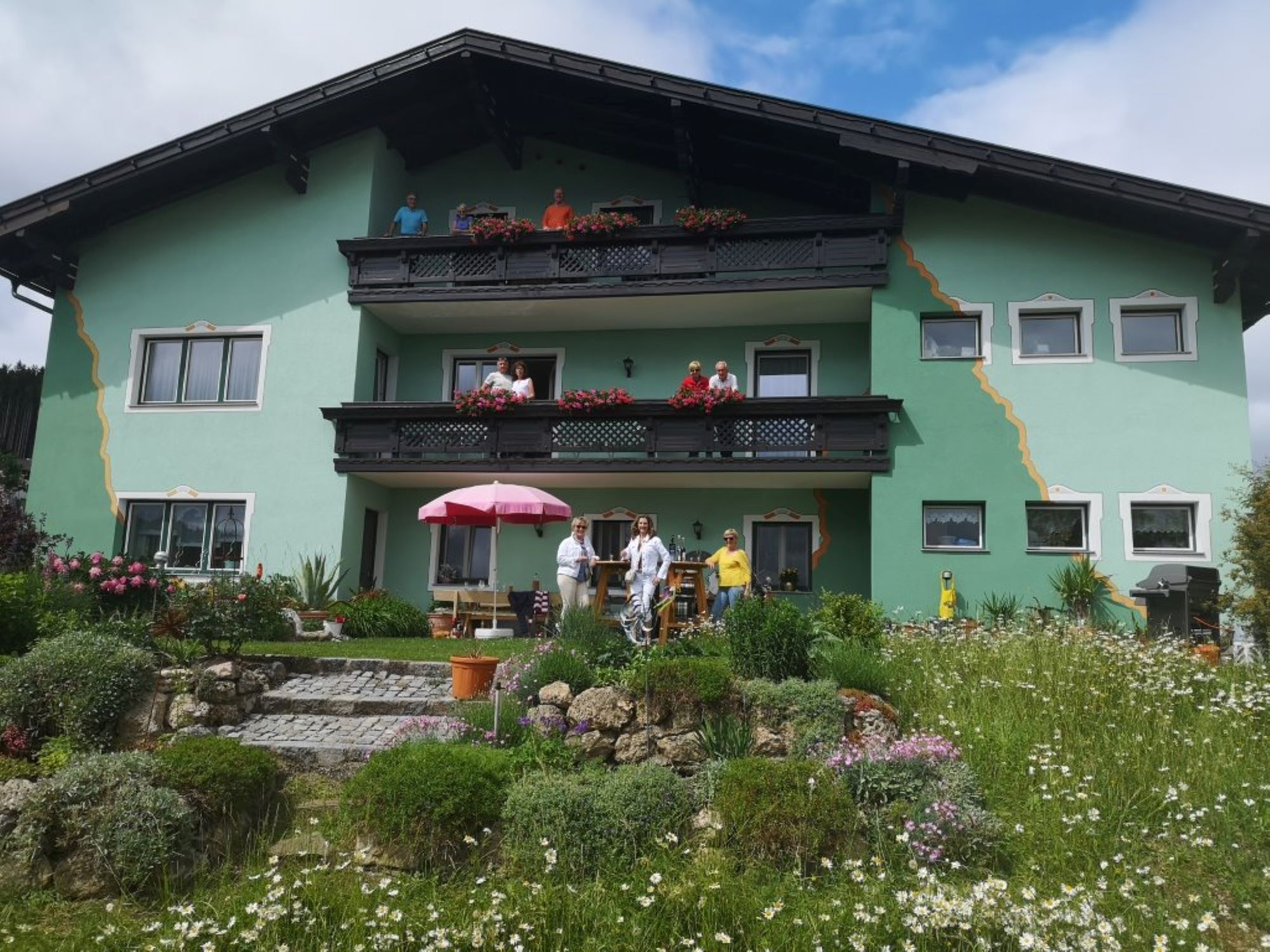 Ein grünes Haus mit Balkonen und Menschen davor, umgeben von einem blühenden Garten.
