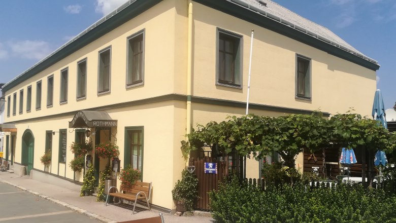 Gelbes Gasthaus mit grünen Fensterrahmen, Gastgarten und Pflanzen vor dem Eingang.