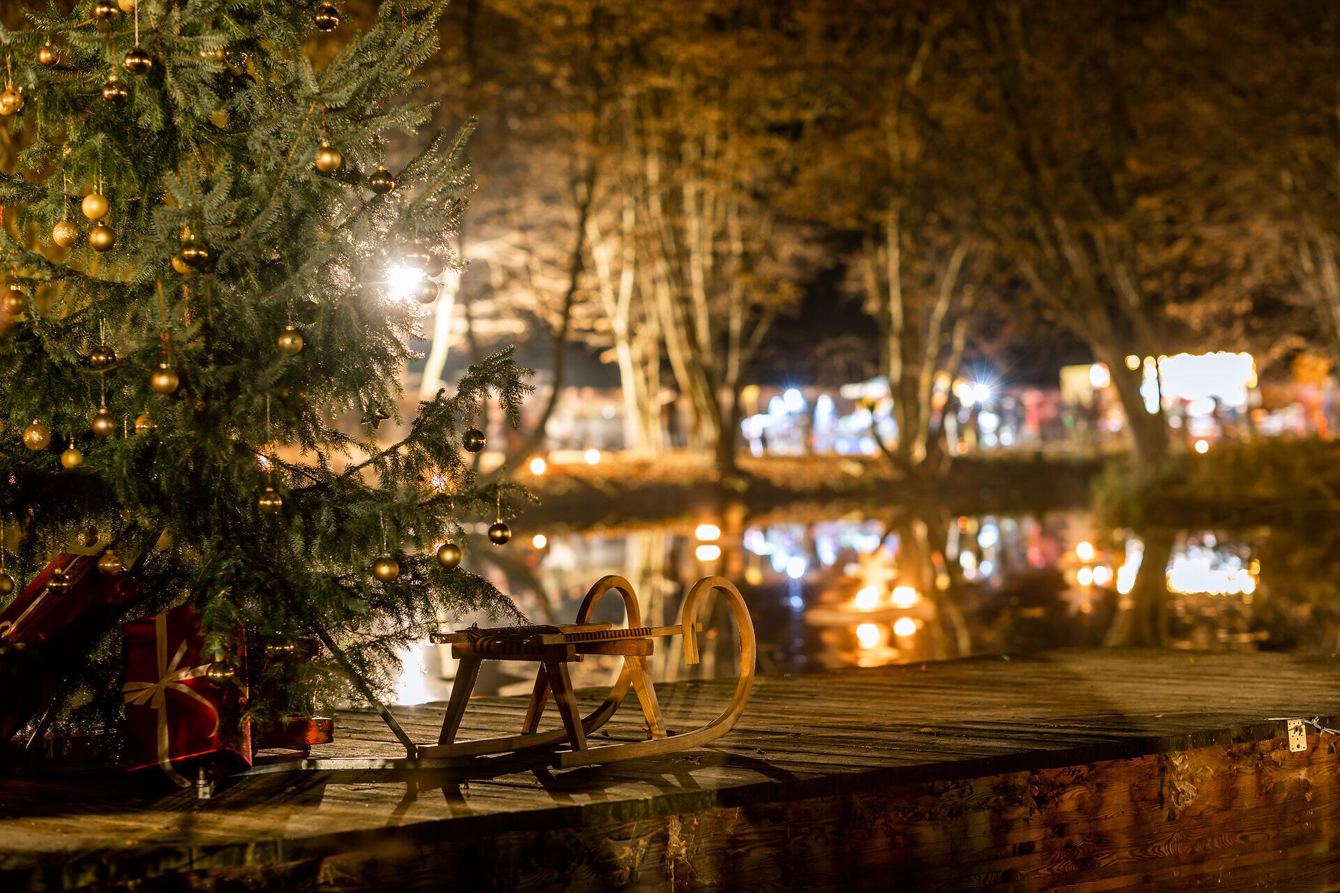 Weihnachtsbaum mit Geschenken und einem Schlitten am Steg eines Teiches mit Spiegelungen von Lichtern in winterlicher Abendstimmung.
