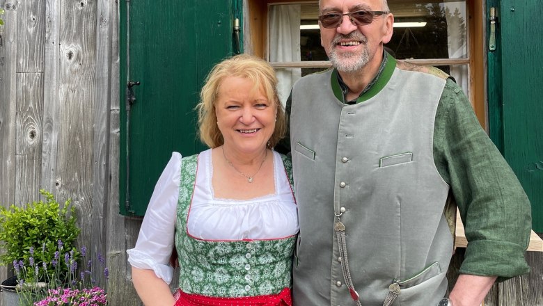 A man and a woman in traditional costume in front of a wooden house with green shutters.