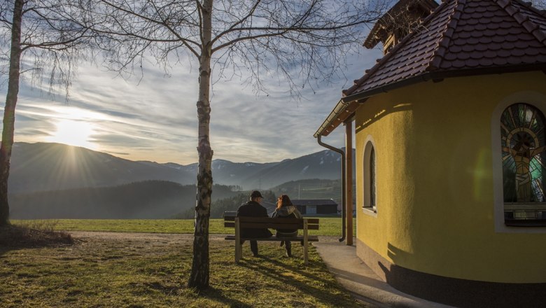 Zwei Personen sitzen auf einer Bank neben einer kleinen Kapelle mit Blick auf die Berge bei Sonnenuntergang.