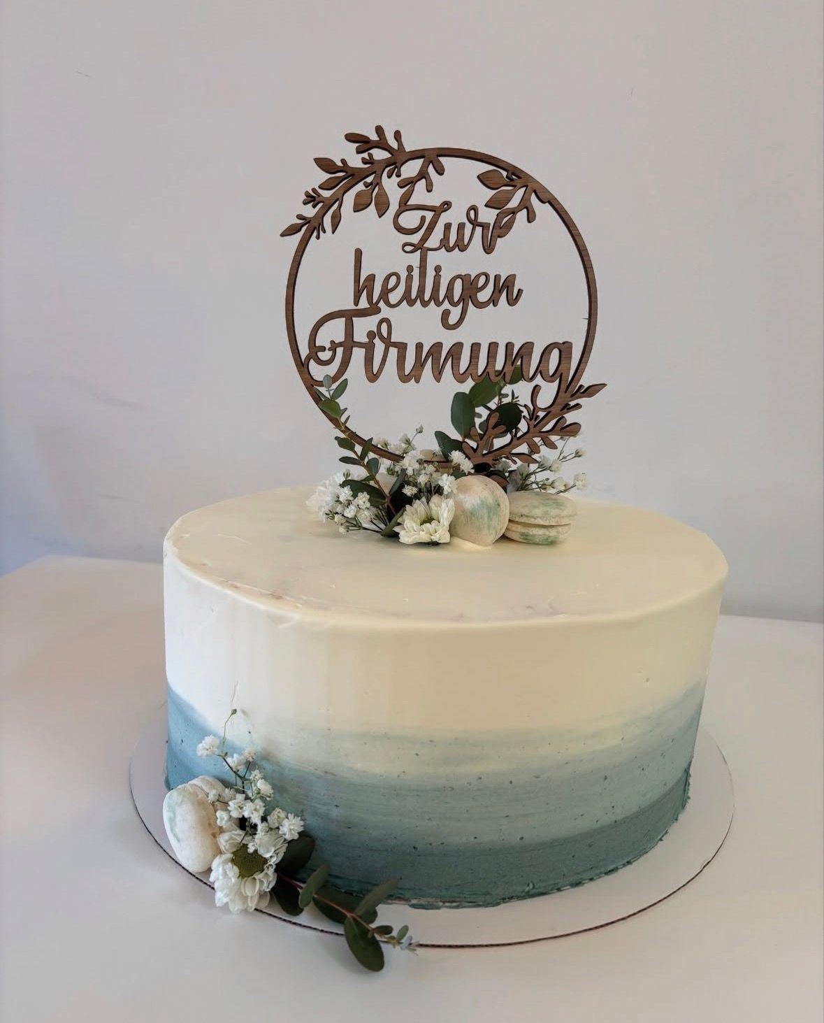 Runde Torte mit weiß-blauem Farbverlauf, Blumen und Macarons; oben ein Holzstecker mit der Aufschrift „Zur heiligen Firmung“.