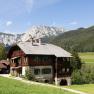 A traditional wooden house in a mountainous landscape with meadows and forests in the background.