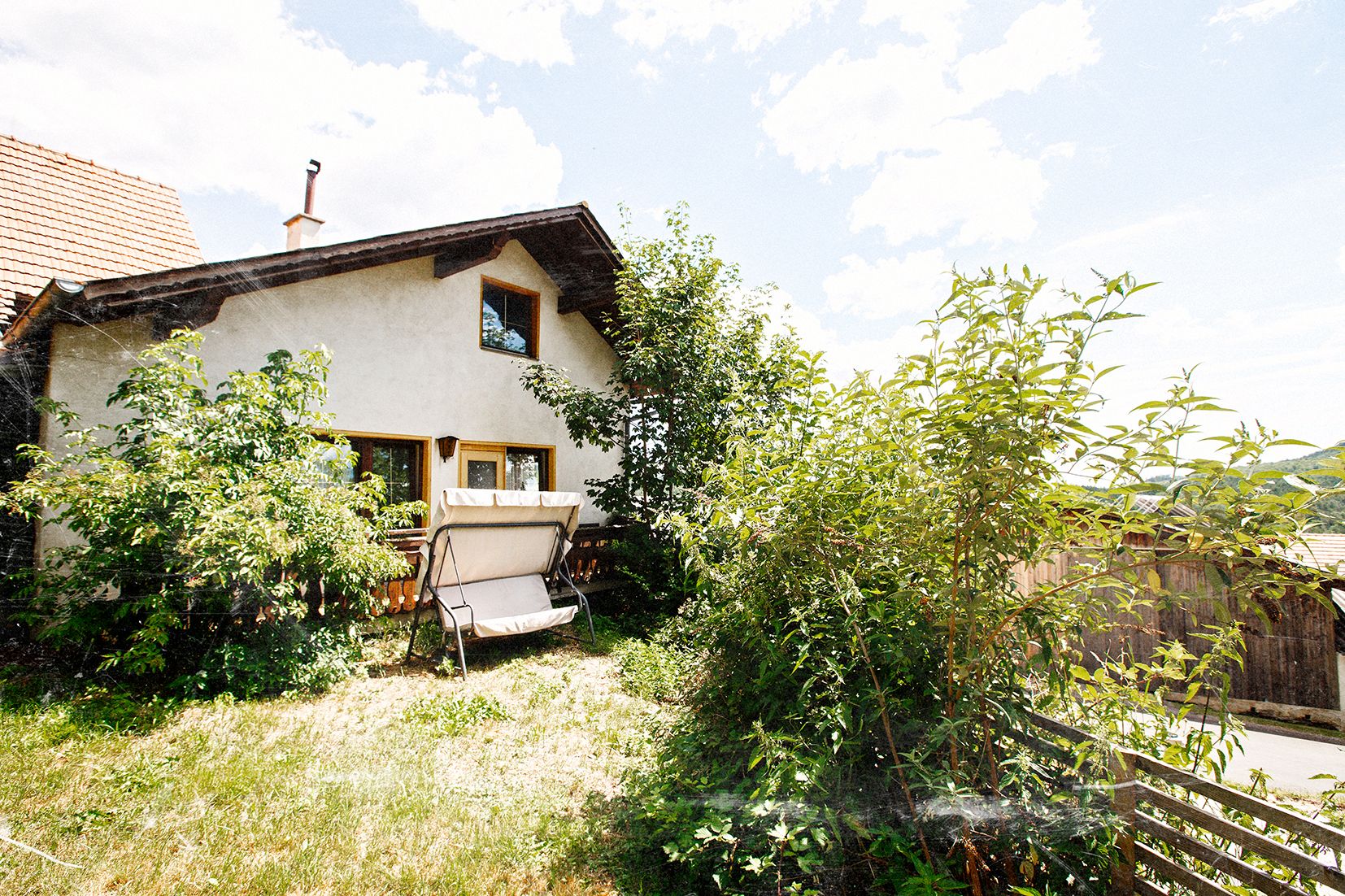 Ein gemütliches Haus mit Garten und Schaukelbank, umgeben von üppigem Grün und Bäumen unter einem blauen Himmel.