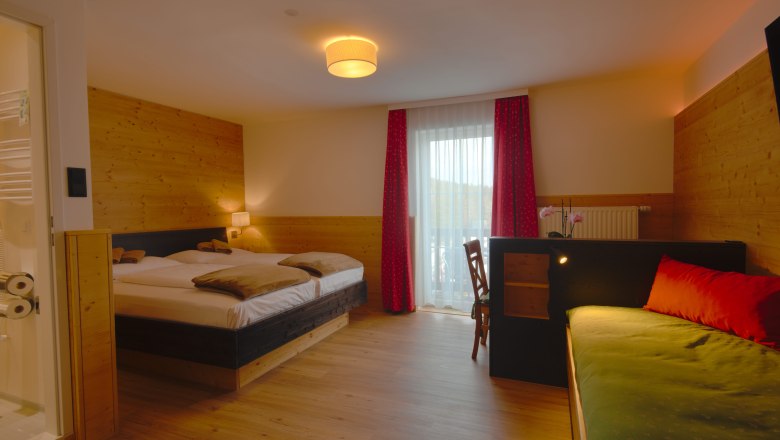 A cozy double room with wooden walls, a double bed, a sofa and a desk. A window with red curtains lets in natural light.