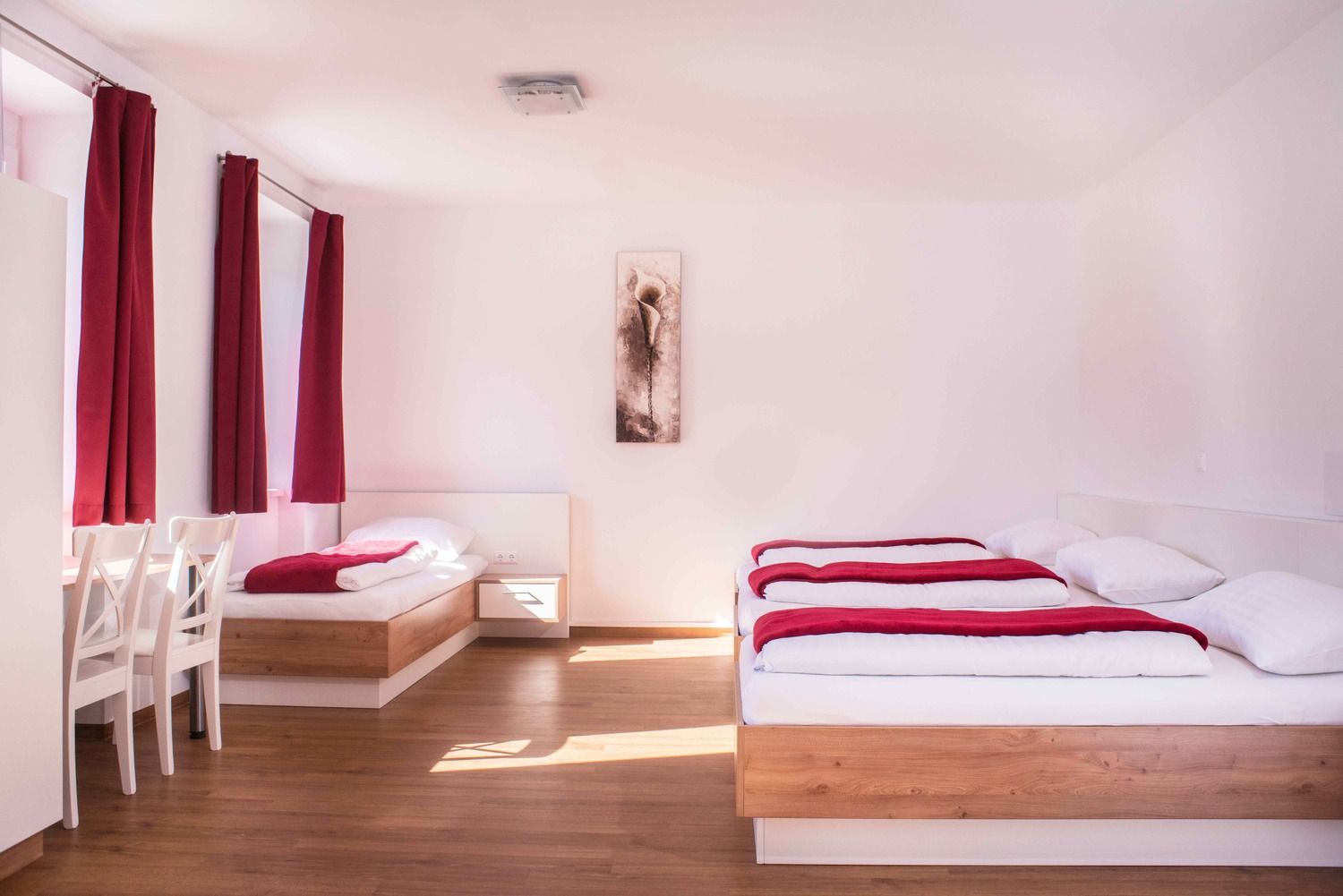 Helles Zimmer mit zwei Betten, roten Vorhängen und einem Bild an der Wand.