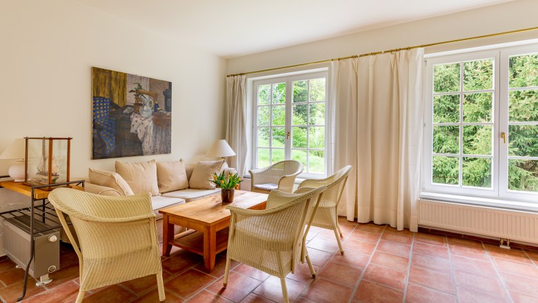 Heller Wohnraum mit Sofa, Korbstühlen, Holztisch und großen Fenstern mit Blick ins Grüne.