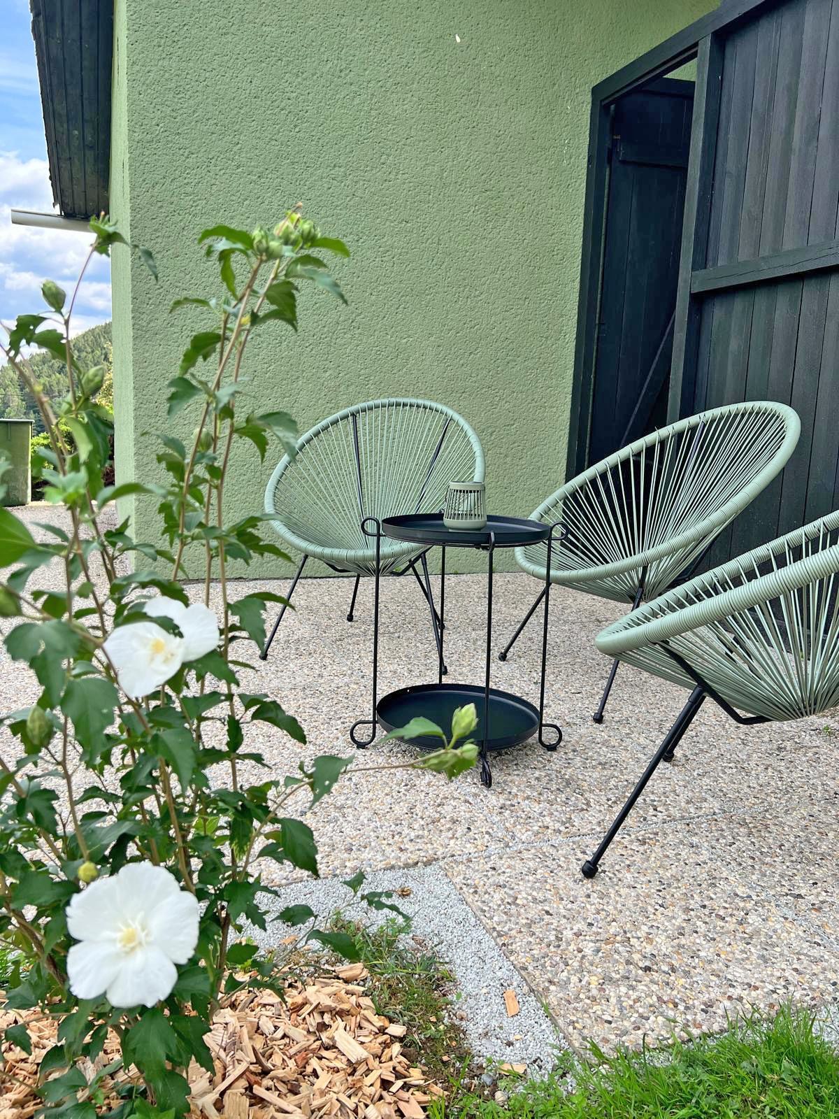 A patio with three mint-green chairs, a black side table, and a house wall painted green; a white shrub is in bloom in the foreground.