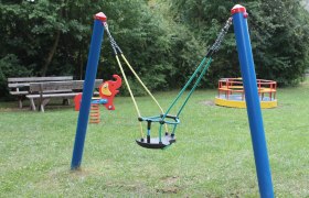 Spielplatz mit Schaukel, Wippe und Karussell auf einer Wiese.