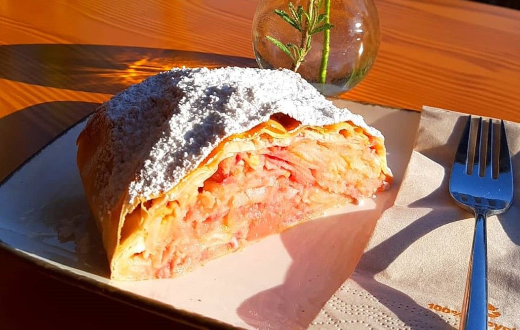 Ein Stück Apfelstrudel mit Puderzucker auf einem Teller, daneben eine Gabel und eine Vase mit Rosmarin.