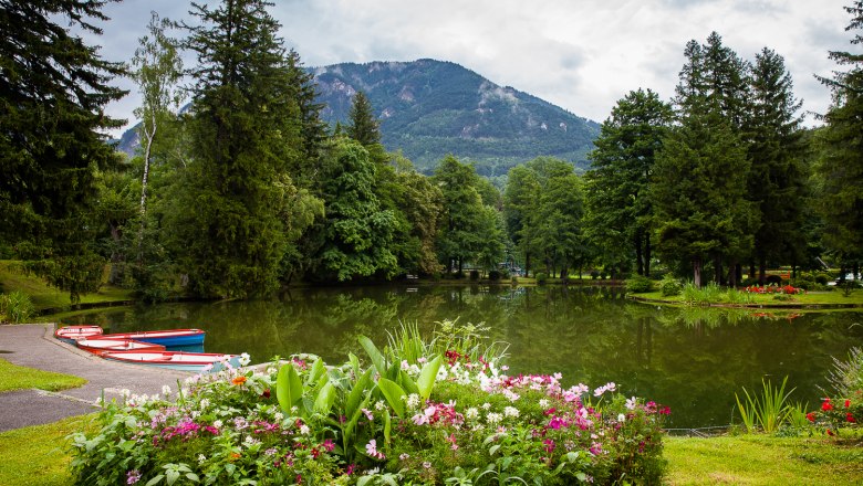 Ein malerischer Park mit einem Teich, umgeben von Bäumen und Blumen, im Hintergrund Berge.