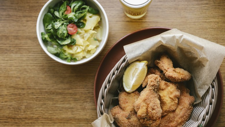 Ein Teller mit Backhendl und Zitronenscheibe, daneben ein Sch&auml;lchen Kartoffel-Vogerlsalat und ein Glas Bier auf einem Holztisch.
