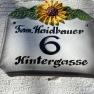 Hausnummernschild mit Sonnenblume und der Aufschrift 'Fam. Haidbauer 6 Hintergasse'.