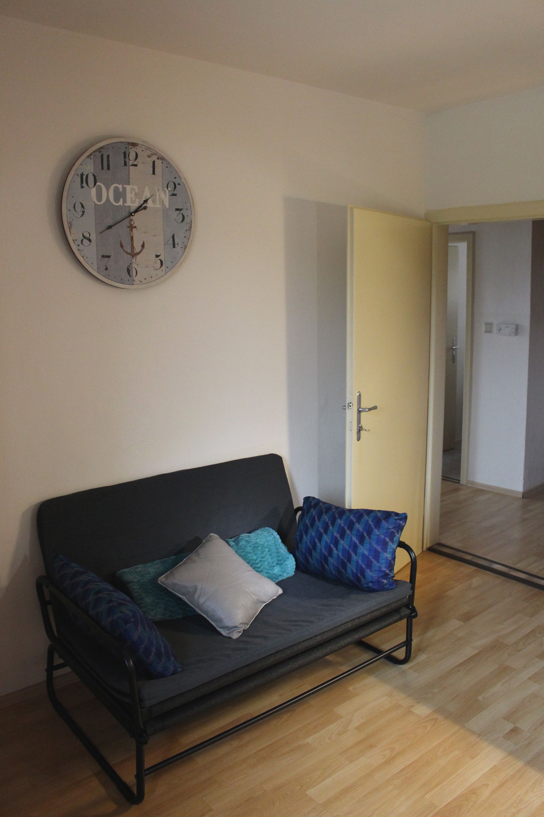 Ein Wohnzimmer mit einer grauen Couch, blauen Kissen und einer Wanduhr mit der Aufschrift 'Ocean'.