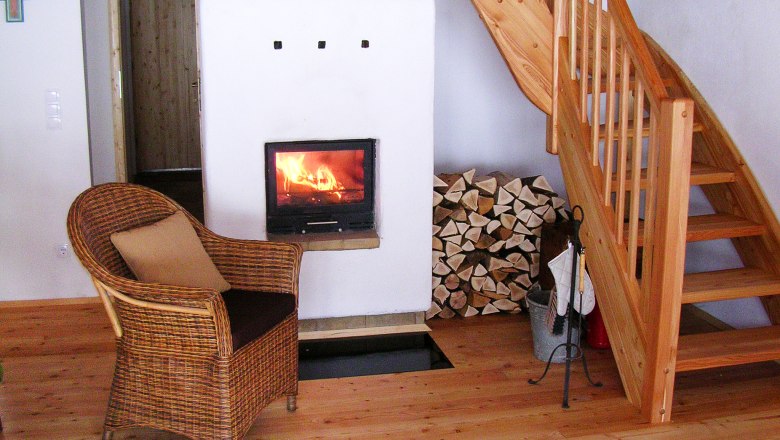 Innenraum eines Ferienhauses mit Kamin, Holzstapel, Korbsessel und Holztreppe.