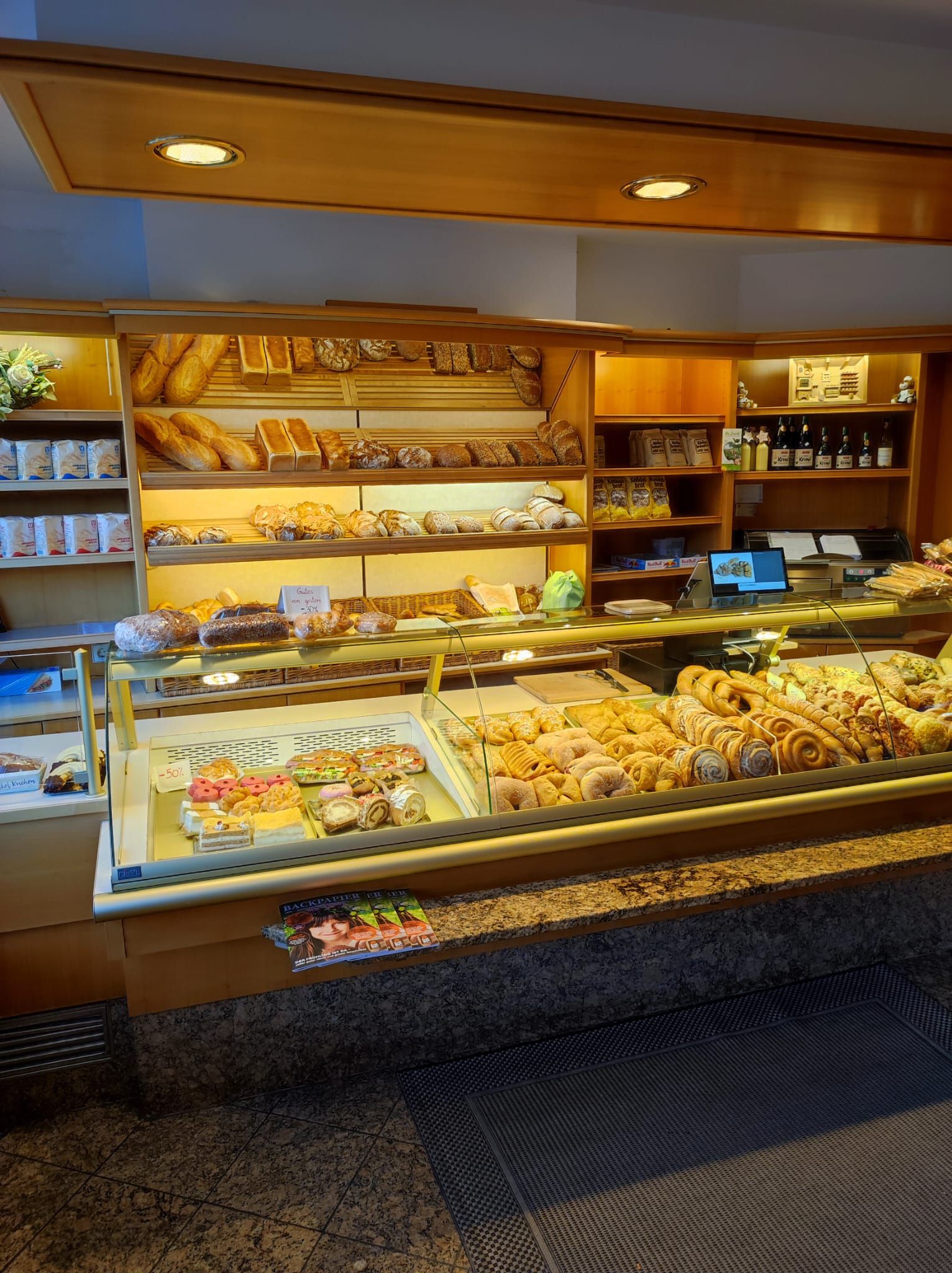 Innenraum von Cafe Dietmayer in Ternitz mit beleuchteter Theke voller Brot, Gebäck und Kuchen sowie Regalen mit Backwaren und Getränken.