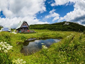 Umgeben von üppigen Wiesen und majestätischen Bergen, lädt die idyllische Hütte zu einer wohlverdienten Rast ein. Die klare Luft und das sanfte Plätschern des Wassers schaffen eine harmonische Atmosphäre, die Wanderer in ihren Bann zieht.