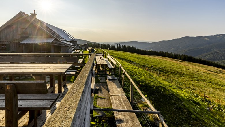 Eine Berghütte mit Holzbänken und Tischen, umgeben von grünen Wiesen und Bergen im Sonnenlicht.