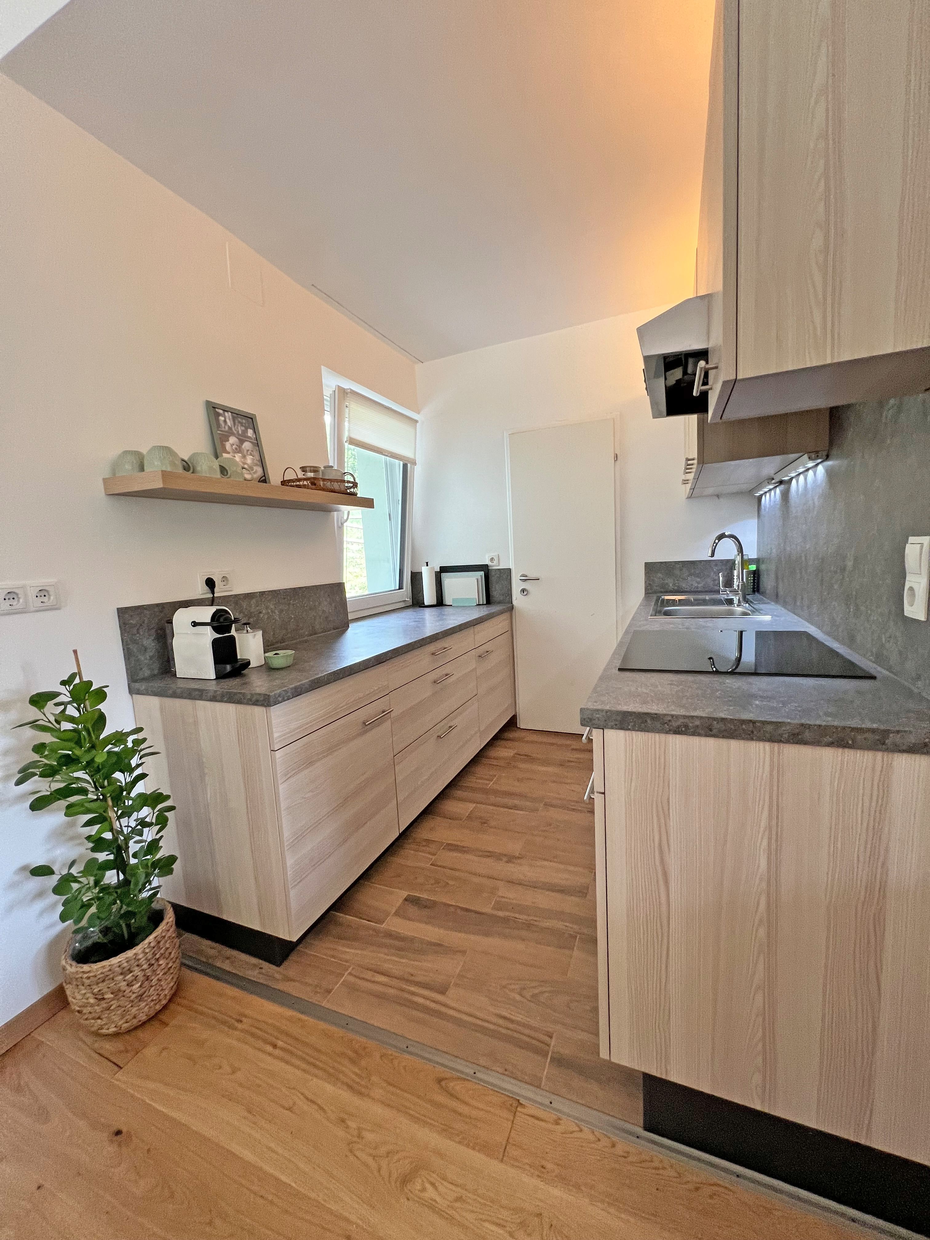 A modern, narrow kitchen with light-colored wood cabinet fronts, gray countertops, a window, a sink, a coffee maker, and a plant in a basket.