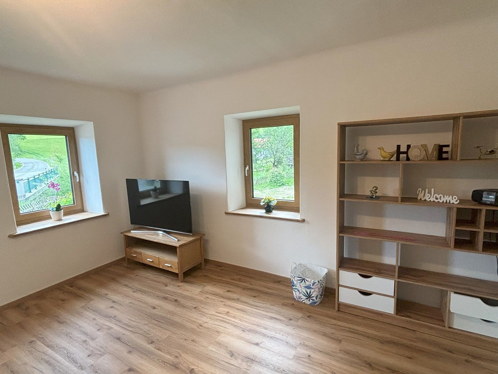 Ein Wohnzimmer mit Holzfußboden, einem Fernseher auf einem Tisch, zwei Fenstern und einem Regal mit Dekorationen.