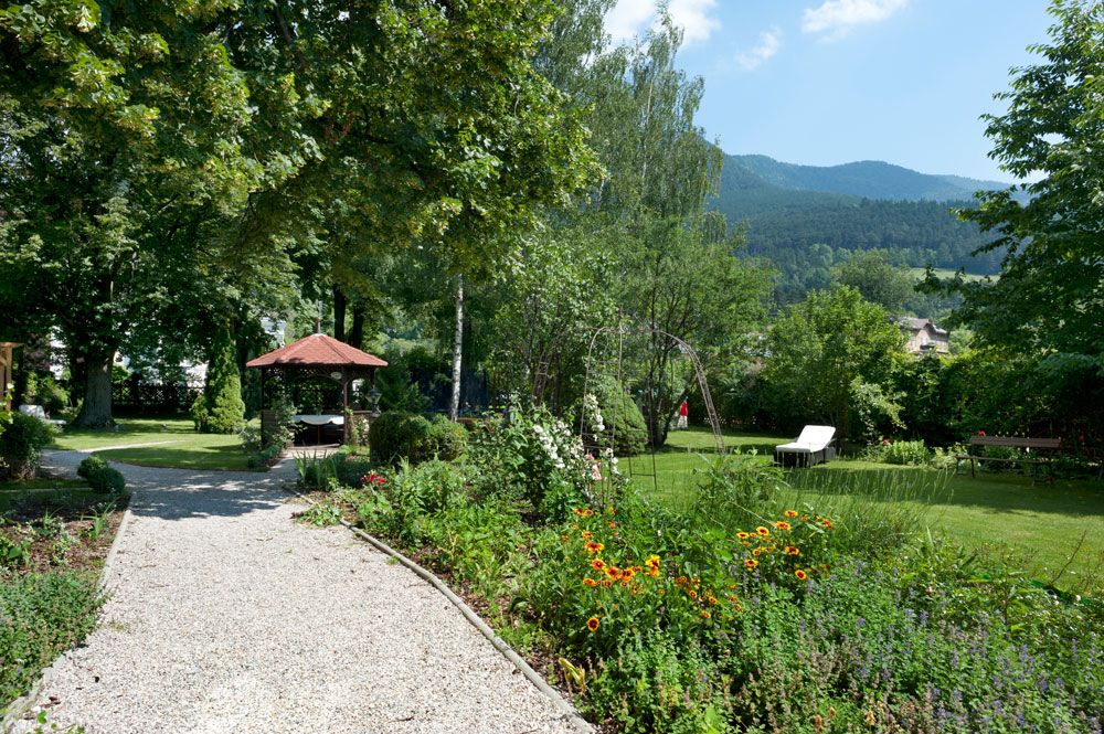 Ein gepflegter Garten mit Kiesweg, Blumenbeeten, einem Pavillon und Liegestühlen vor einer Bergkulisse.