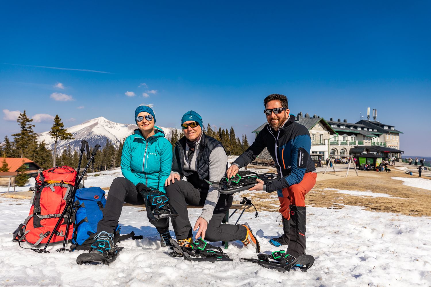 Drei Personen mit Schneeschuhen sitzen und stehen im Schnee vor der Raxalpe, mit Berghütte und blauem Himmel im Hintergrund.