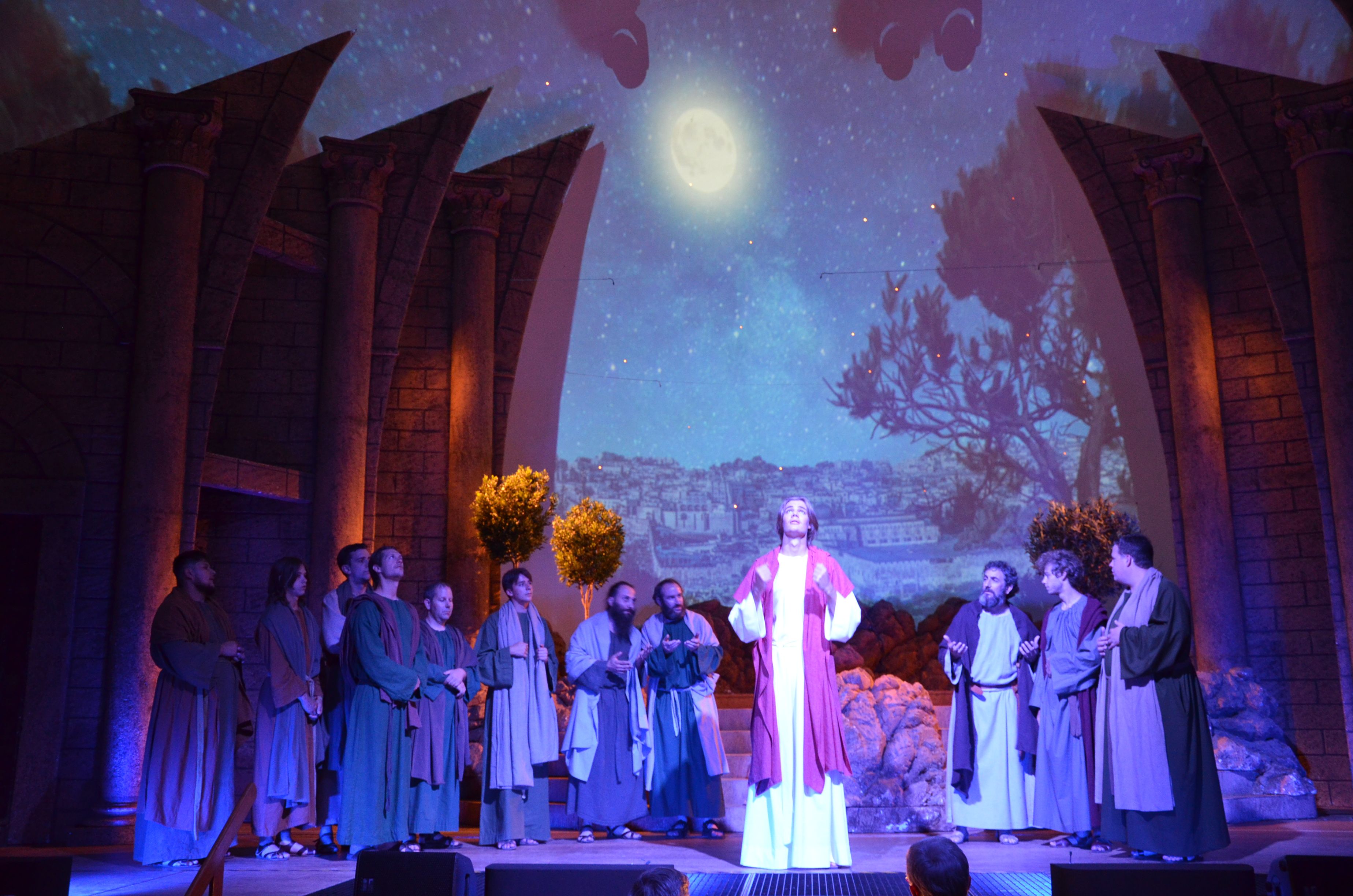 Szene aus den Passionsspielen: Jesus steht in einer Lichtung, die Lichtstimmung symbolisiert den Abend, der Mond wird auf das Bühnenbild projiziert. Um ihn haben sich Menschen versammelt.