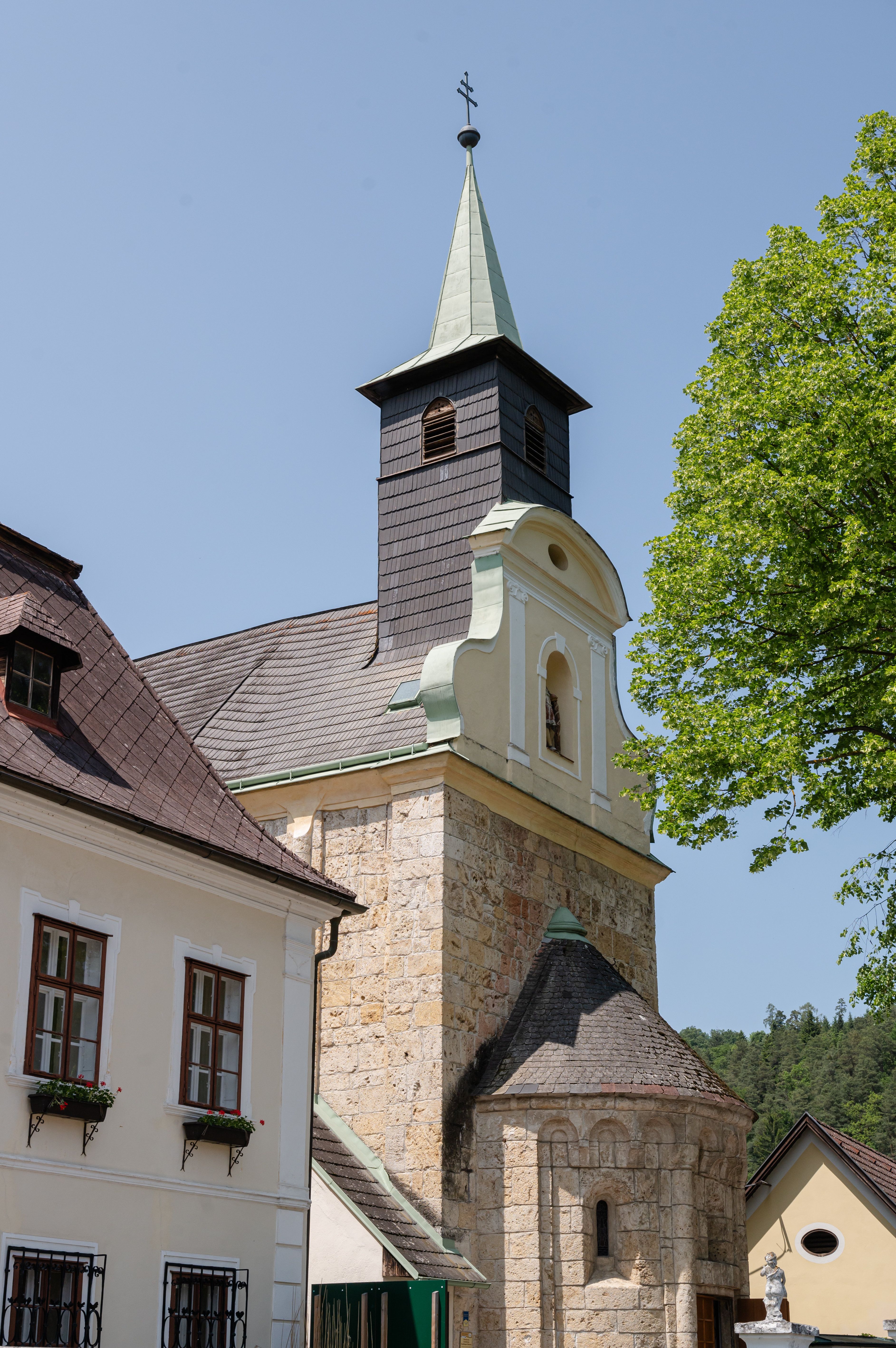Kirchturm mit grünem Dach und Kreuz vor blauem Himmel, umgeben von Gebäuden und Bäumen in Thernberg