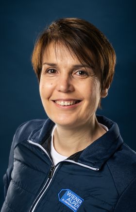 Portraitfoto Wiener Alpen Mitarbeiterin, die eine Jacke mit Wiener Alpen Logo trägt und vor einem dunklem Hintergrund steht.