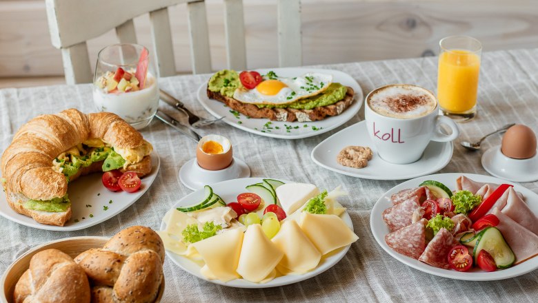 Ein reichhaltiges Frühstück mit Croissant, Brötchen, Käse, Aufschnitt, Avocado-Toast, Joghurt, Cappuccino, Orangensaft und weichen Eiern auf einem Tisch.