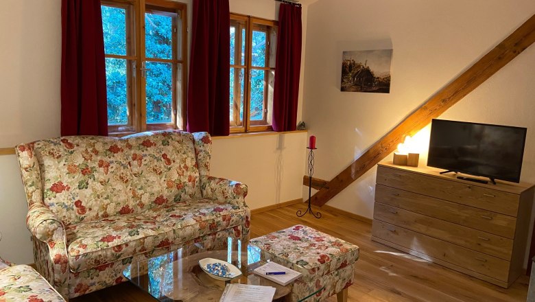 Gem&uuml;tliches Wohnzimmer mit floralen Sofas, Holzm&ouml;beln und einem Fernseher. Rote Vorh&auml;nge und ein Bild an der Wand.