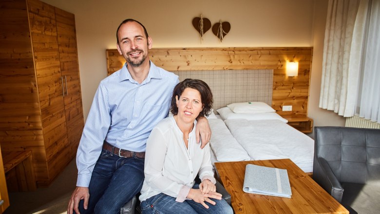 Ein Paar sitzt in einem gem&uuml;tlichen Zimmer mit Holzm&ouml;beln und einem Doppelbett.