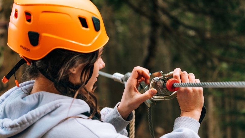 Person mit orangefarbenem Helm befestigt Sicherheitsausrüstung an einem Seil im Waldseilgarten.