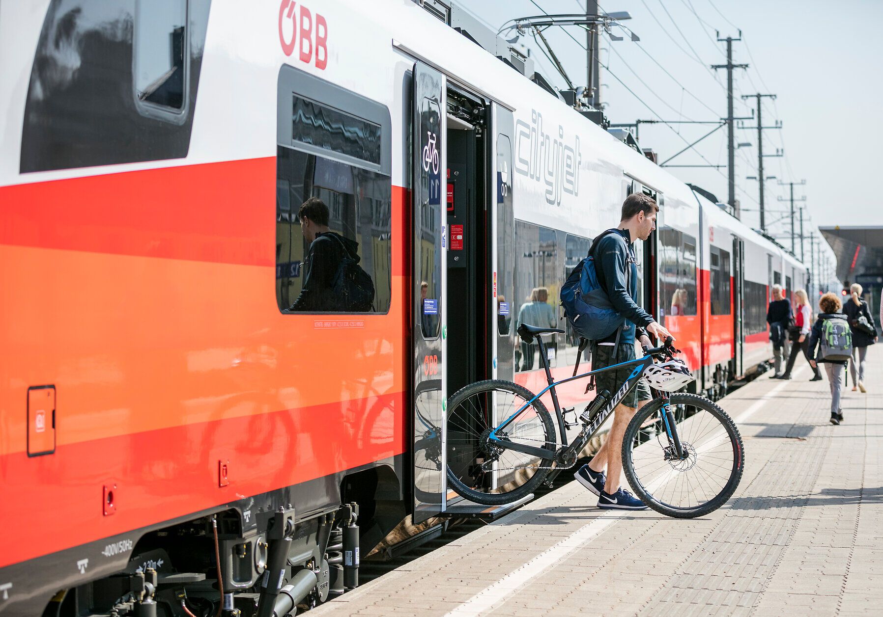 Die Sonne strahlt über den Bahnhof, während Reisende mit ihren Fahrrädern in den Zug einsteigen. Die frische Luft und die Vorfreude auf neue Abenteuer in der Natur sind spürbar. Ein perfekter Start für eine unvergessliche Reise in die Berge.
