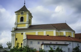 Gelbe Kirche mit Turm und roten Dächern vor bewölktem Himmel.