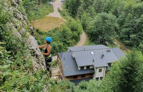 Person climbing on a rock face with helmet and climbing equipment, buildings and forest in the background.