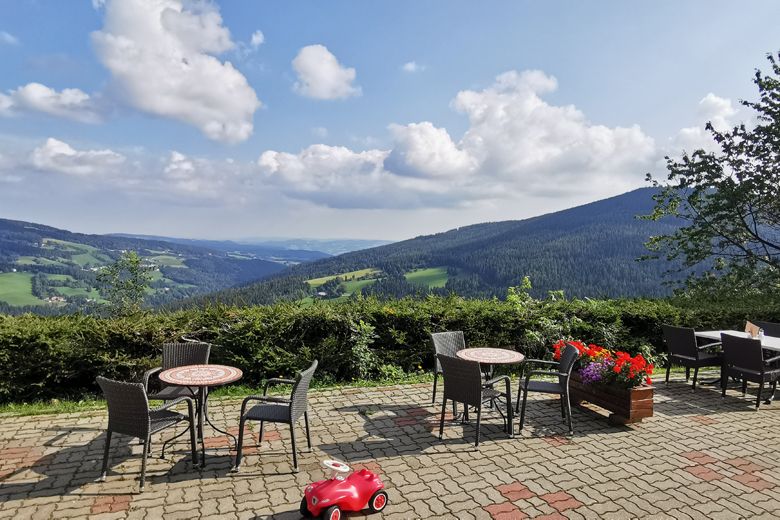 Terrasse mit Tischen und Stühlen, Blick auf grüne Hügel und blauen Himmel mit Wolken.