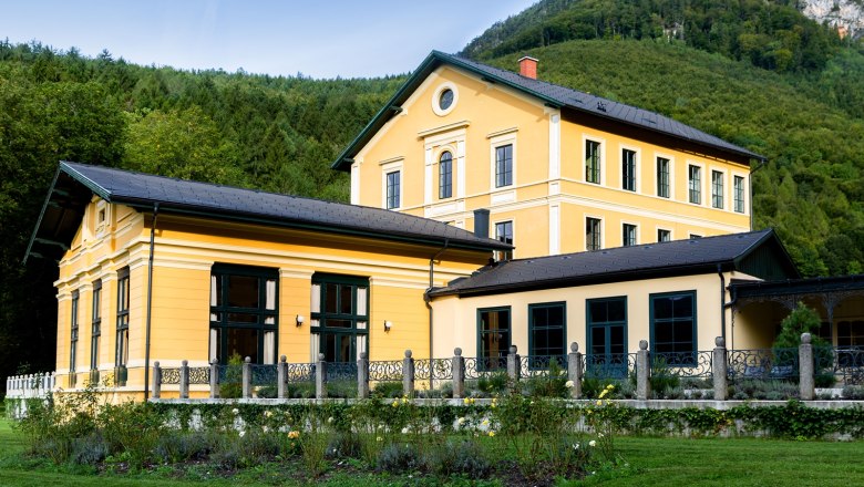 Au&szlig;enansicht eines gelben Geb&auml;udes mit Garten und Wald im Hintergrund.