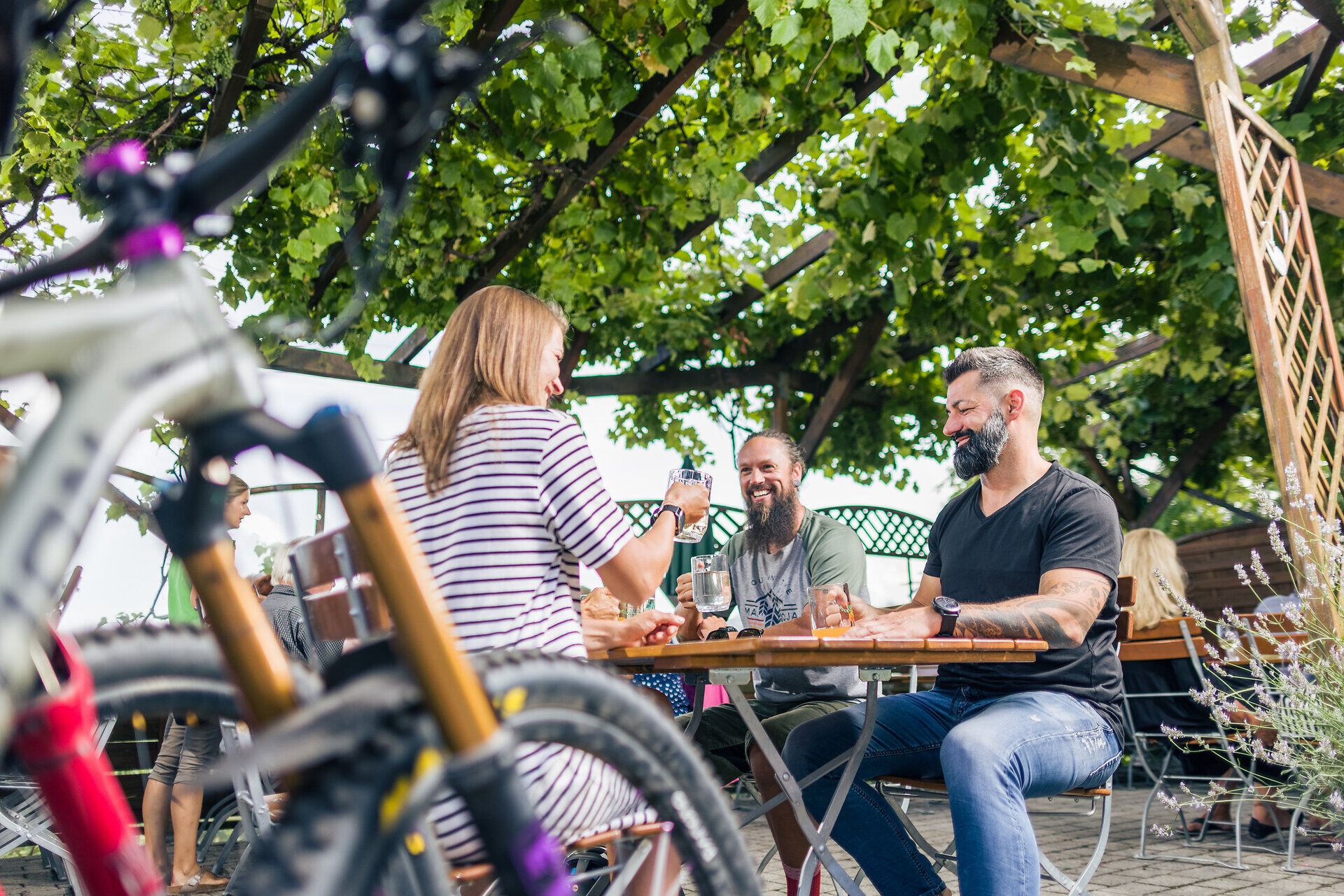Eine Frau und zwei Männer sitzen lachend mit Gläsern in ihren Händen an einem Tisch unter einer mit Weinrebe bedeckten Holzpergola. Im Vordergrund steht ein Fahrrad.