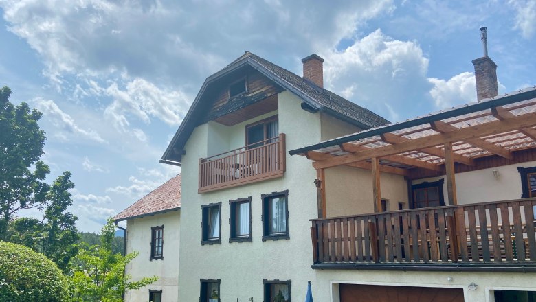 Ferienwohnungen Riedl, © Wiener Alpen Ein zweistöckiges Haus mit Balkon und Terrasse, umgeben von Garten und Bäumen, unter einem bewölkten Himmel.